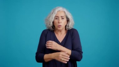 Studio video with blue background of an aged caucasian woman looking at camera while embracing herself expressing coldness