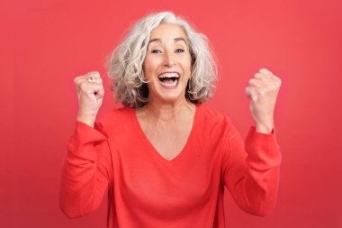 Studio portrait with red background of a mature woman celebrating while raising the fists