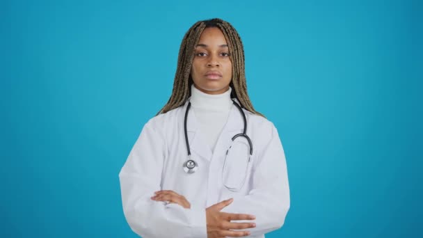 青い背景を持つビデオの深刻なアフリカの女性医師とともに腕を組んでカメラを見て — ストック動画