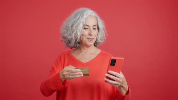 携帯電話でオンラインショッピングにクレジットカードを使用して成熟した女性の赤い背景を持つスタジオビデオ — ストック動画