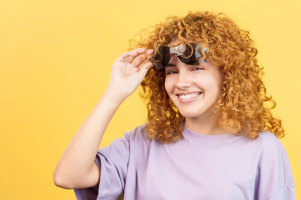 Studio image with yellow background of a cool woman with curly hair lifting up her sunglasses and looking at the camera