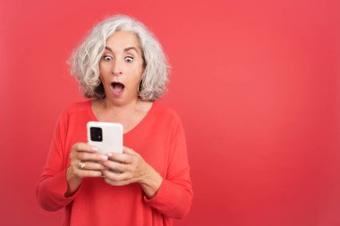 Studio portrait with red background of a surprised mature woman using the mobile phone