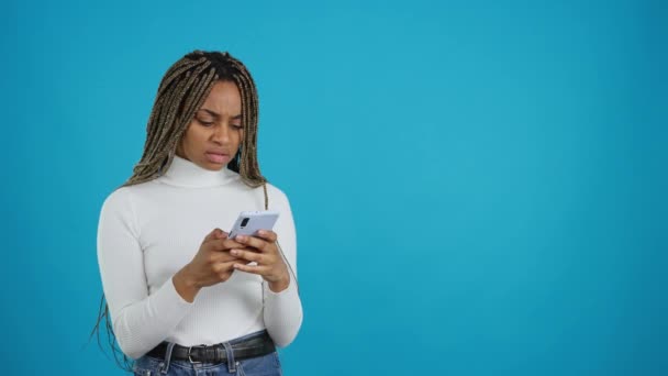 怒りと携帯電話を見て信じられないほどの表現を持つアフリカの女性の青の背景を持つビデオ — ストック動画