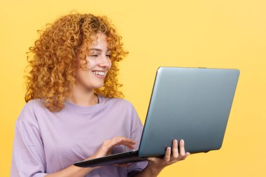 Studio image with yellow background of a beauty and smiley woman with curly hair standing and using a laptop