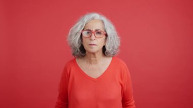 Studio video with red background of a mature woman lifting up her glasses to looking at camera