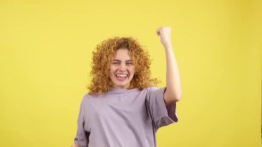 Studio video with yellow background of a beauty woman with curly hair looking at camera while celebrating raising the fists