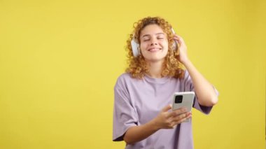 Studio video with yellow background of a smiley woman with curly hair listening to music with a mobile and headphones