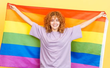 Studio image with yellow background of a beauty and happy woman with curly hair waving a lgbt rainbow flag