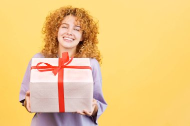 Studio image with yellow background of a happy young woman smiling at camera while giving a gift