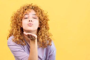 Studio image with yellow background of a young woman with curly hair blowing a kiss to the camera