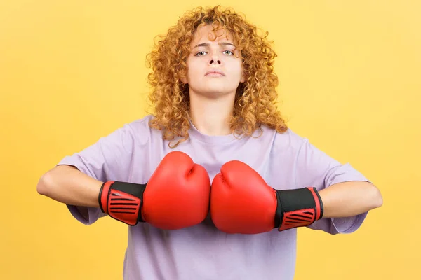 Studio image with yellow background of a young rude woman with curly hair in boxing gloves looking at camera