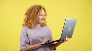 Studio video with yellow background of a beauty and smiley woman with curly hair standing and using a laptop