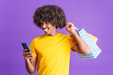 Smiley african man with curly hair using the mobile while holing shopping bags in studio