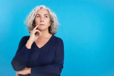 Studio portrait with blue background of an aged woman gesturing and looking up with thoughtful expression