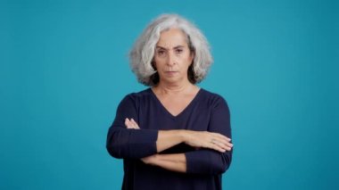 Studio video with blue background of a mature woman crossing the arms while looking at camera with an angry expression