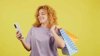 Studio video with yellow background of a happy young woman with curly hair taking a selfie while holing shopping bags