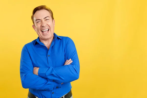 Mature man laughing while standing with arms crossed in studio with yellow background