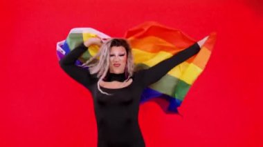 Stüdyoda kırmızı arka planda lgbt gökkuşağı bayrağı taşıyan travesti kılığında mutlu bir transseksüel.