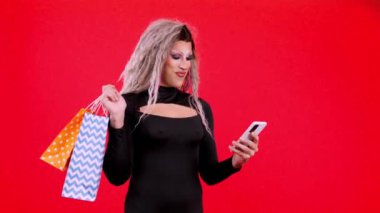transgender person dressed as drag queen using a mobile while holding shopping bags in studio with red background
