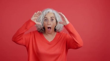 Studio video with red background of a mature woman gesturing with a surprised face with the mouth opened