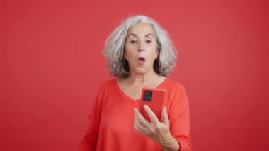 Studio video with red background of a mature woman celebrating while using the mobile phone