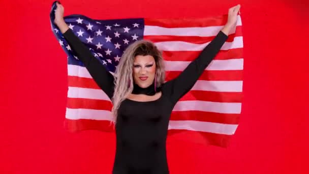 幸せなトランスジェンダーの人は 赤の背景を持つスタジオで北アメリカの国旗を掲揚するドラッグクイーンとして身に着けています — ストック動画