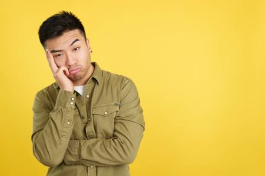 Bored chinese man standing and looking at camera in studio with yellow background