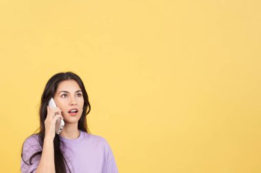 Surprised hispanic woman talking to the mobile in studio with yellow background