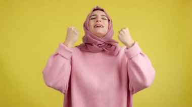 Happy muslim woman celebrating while raising fists in studio with yellow background
