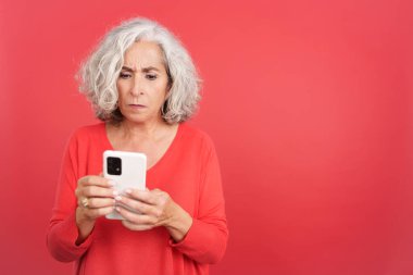 Studio portrait with red background of a worried mature woman using a mobile phone