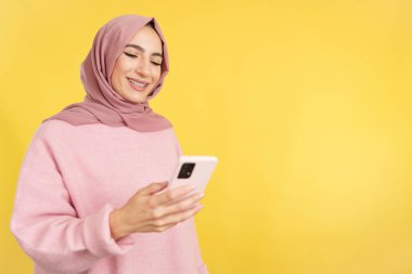 Happy muslim woman smiling while using a mobile phone in studio with yellow background