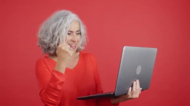 Yetişkin bir kadının dizüstü bilgisayar kullanırken kutlama yaptığı kırmızı arka planlı bir stüdyo videosu