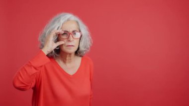 Studio portrait with red background of a mature woman lifting up her glasses to see from a distance