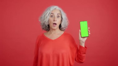 Studio video with red background of a surprised mature woman showing the chroma screen of the mobile, Mock up
