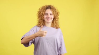 Studio video with yellow background of a happy young woman with curly hair gesturing agreement raising the thumb