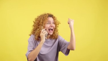 Studio video with yellow background of an excited woman with curly hair celebrating and screaming while taking to the mobile