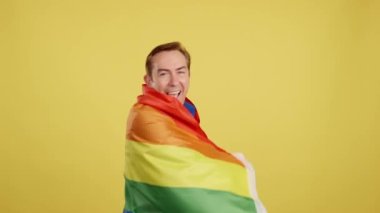 Happy mature man wrapped with a lgbt flag in studio with yellow background