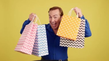 Smiley mature man looking at the camera showing shopping bags in studio with yellow background