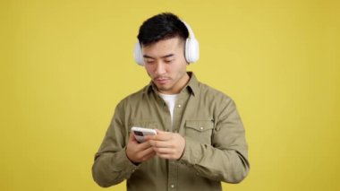 Happy chinese man dancing and listening to music with the mobile phone and headphones in studio with yellow background