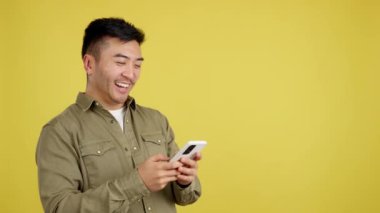 Stüdyoda sarı arka planda cep telefonunu kullanırken gülümseyen mutlu Çinli adam.