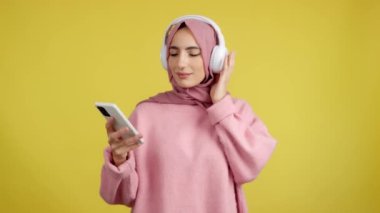 Happy muslim woman smiling while listening to music with the mobile and headphones in studio with yellow background