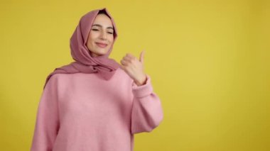 Muslim woman smiling at camera and pointing to the side in studio with yellow background