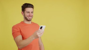 Happy caucasian man smiling while using the mobile in studio with yellow background