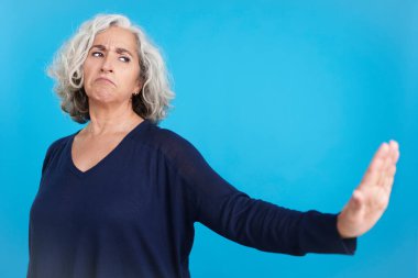 Studio portrait with blue background of a mature woman gesturing that she wants to know nothing by pushing away with her hand