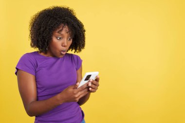 Surprised woman with afro hair using a mobile in studio with yellow background