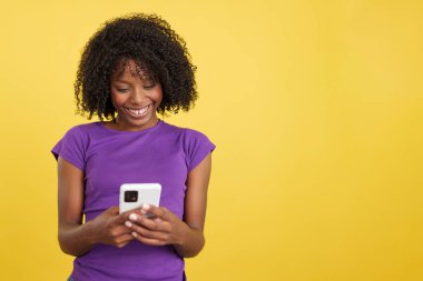 Woman with afro hair smiling while sending a message with a mobile in studio with yellow background