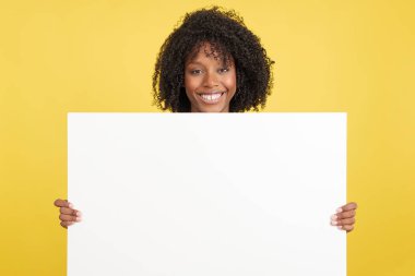 Woman with afro hair holding a blank panel while smiling in studio with yellow background