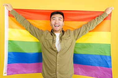 Happy chinese man raising a lgbt rainbow flag in studio with yellow background