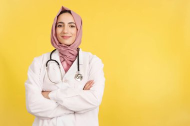 Happy female muslim doctor with arms crossed looking at camera in studio with yellow background