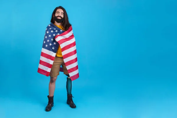Studio portrait with blue background of a man with prosthetic leg wrapped in a north america national flag
